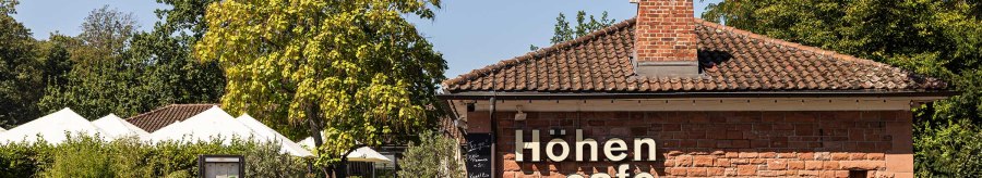 The H&ouml;hencaf&eacute; in Killesbergpark is a brick building with a terrace surrounded by trees and plants. White parasols are visible., &copy; Stuttgart-Marketing GmbH, Sarah Schmid