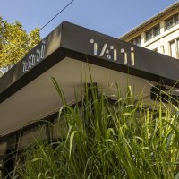 Modern building with the lettering 'Tatti' on the roof. Tall green plants can be seen in the foreground. A historic building in the background., © SMG, Sarah Schmid Modern building with the lettering 'Tatti' on the roof. Tall green plants can be seen in the foreground. A historic building in the background., © SMG, Sarah Schmid