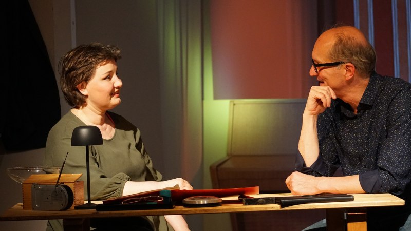 Zwei Personen sitzen an einem Tisch und unterhalten sich in einem warm beleuchteten Raum. Auf dem Tisch liegen verschiedene Gegenstände., © Katja Ritter Zwei Personen sitzen an einem Tisch und unterhalten sich in einem warm beleuchteten Raum. Auf dem Tisch liegen verschiedene Gegenstände., © Katja Ritter