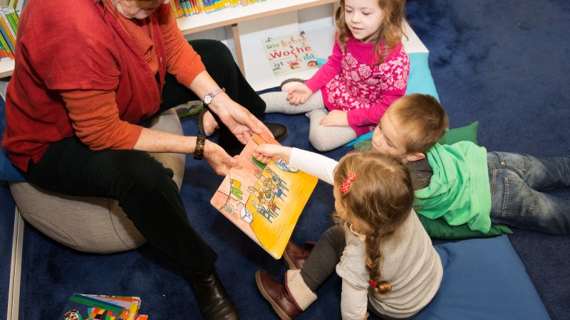 Eine Frau liest drei Kindern in einer Bibliothek ein Bilderbuch vor. Die Kinder sitzen auf Kissen und hören aufmerksam zu., © Stadtbibliothek Stuttgart - Leseohren e.V. Eine Frau liest drei Kindern in einer Bibliothek ein Bilderbuch vor. Die Kinder sitzen auf Kissen und hören aufmerksam zu., © Stadtbibliothek Stuttgart - Leseohren e.V.