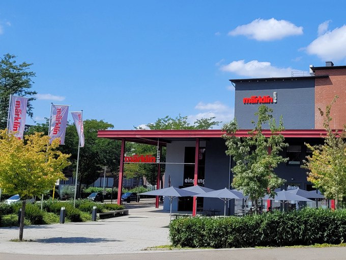 The Märklineum building with red lettering and entrance, surrounded by trees and flags. The sky is blue with a few clouds., © Märklin Spielwaren Vertriebs-GmbH The Märklineum building with red lettering and entrance, surrounded by trees and flags. The sky is blue with a few clouds., © Märklin Spielwaren Vertriebs-GmbH