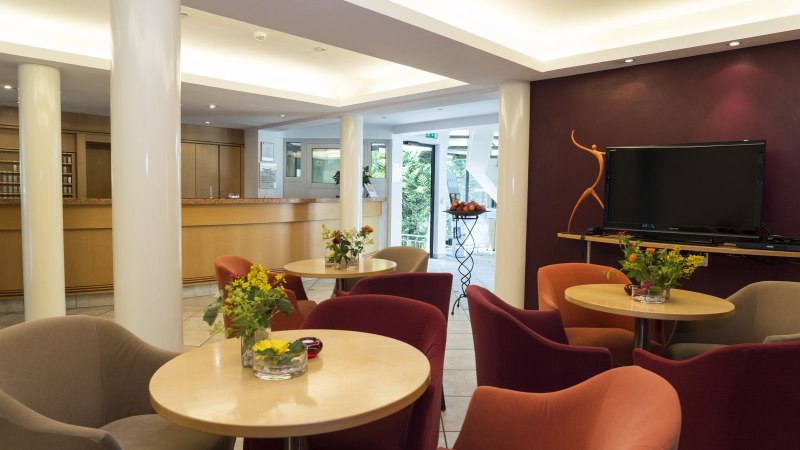 Modern hotel lobby with cozy armchairs, tables with flowers, a large TV and a reception desk in the background., © Hotel Gloria GmbH Restaurant Möhringer Hexle Modern hotel lobby with cozy armchairs, tables with flowers, a large TV and a reception desk in the background., © Hotel Gloria GmbH Restaurant Möhringer Hexle