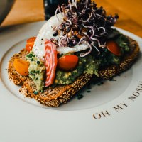 Belegtes Brot mit Avocado, Tomaten und Sprossen auf einem Teller mit der Aufschrift 'Oh my Nosh'., © The Gardeners Nosh Belegtes Brot mit Avocado, Tomaten und Sprossen auf einem Teller mit der Aufschrift 'Oh my Nosh'., © The Gardeners Nosh