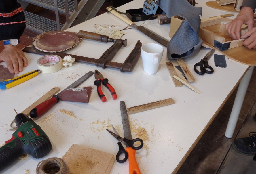 Werkbank mit verschiedenen Werkzeugen wie Bohrer, Zangen, Scheren und Holzstücken in einer Holzwerkstatt. Zwei Personen arbeiten an Projekten., © KVWH