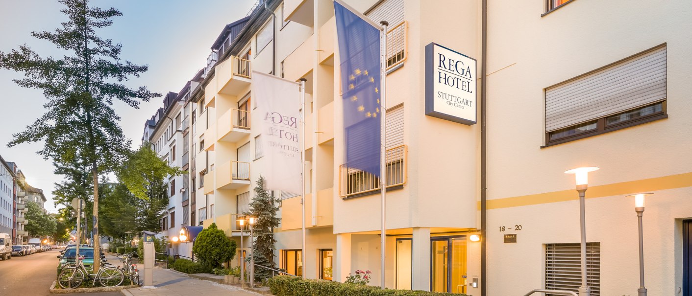 Exterior view of the Rega Hotel in Stuttgart. Multi-storey building with flags and illuminated street lamps. Quiet street with parked cars., © TOMAS