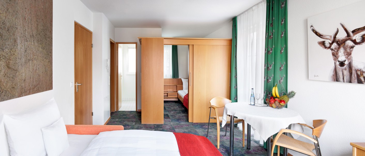 Helles Hotelzimmer mit Doppelbett, Tisch und Stühlen, Holzmöbeln und einem Bild von einem Hirsch an der Wand., © Hirsch Hotel