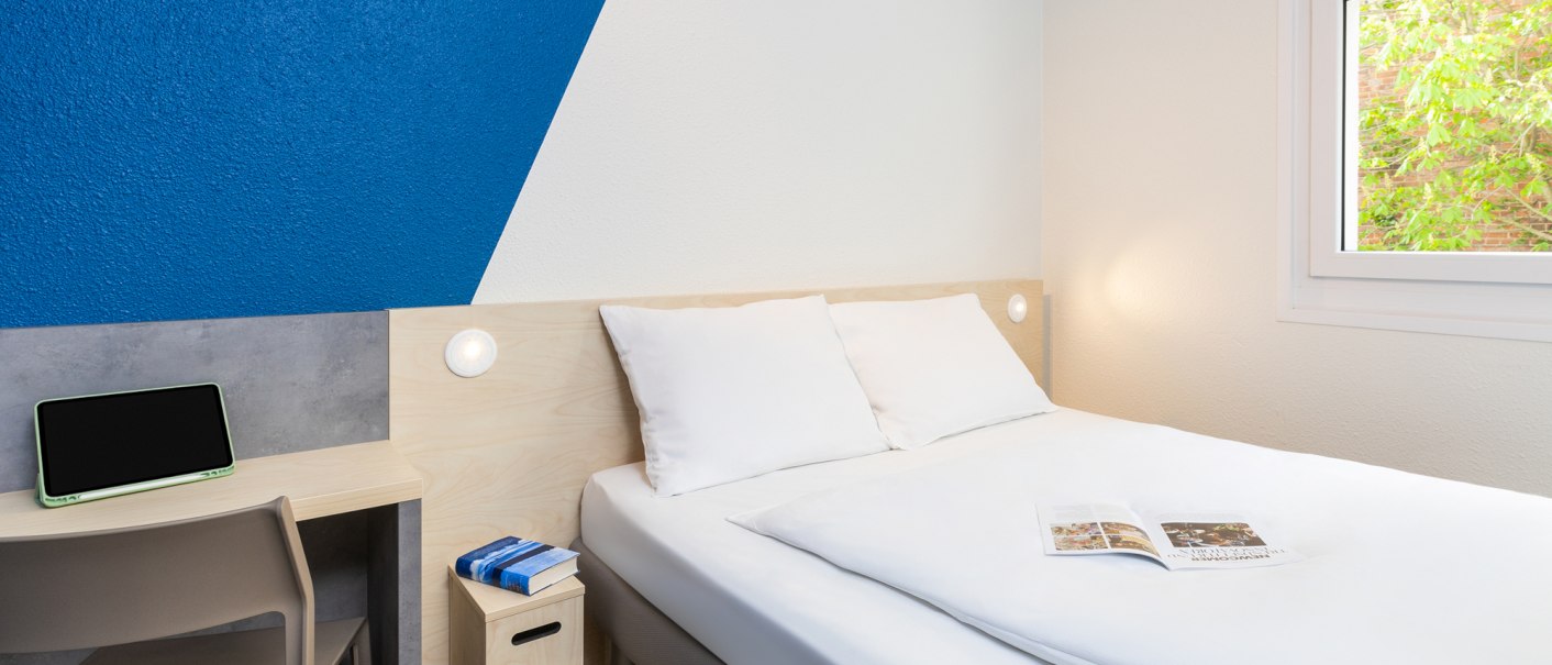Ein schlichtes Hotelzimmer mit Doppelbett, blau-weißer Wand, kleinem Schreibtisch und Fenster mit Blick ins Grüne., © DQuadrat Living GmbH Ein schlichtes Hotelzimmer mit Doppelbett, blau-weißer Wand, kleinem Schreibtisch und Fenster mit Blick ins Grüne., © DQuadrat Living GmbH