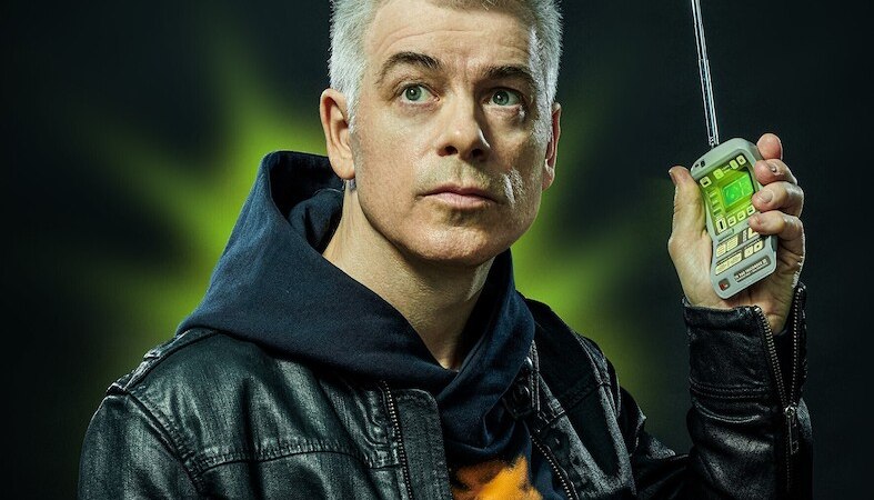 Person mit grauen Haaren in Lederjacke h&auml;lt ein Retro-Handy mit Antenne. Hintergrund ist dunkel mit gr&uuml;nem Lichtschein., &copy; Theaterhaus Stuttgart e.V.