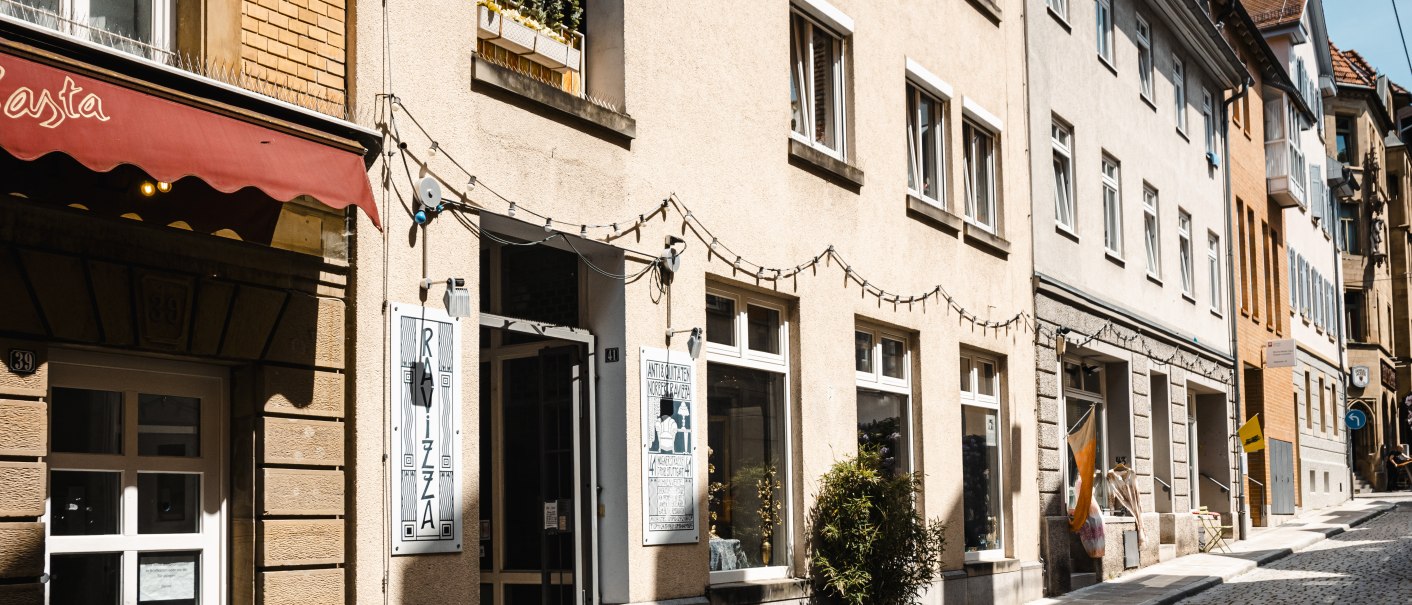 Eine sonnige Gasse mit Kopfsteinpflaster, gesäumt von Geschäften wie einem Antiquariat. Lichterketten hängen über den Eingängen., © SMG Stuttgart Marketing GmbH - Sarah Schmid