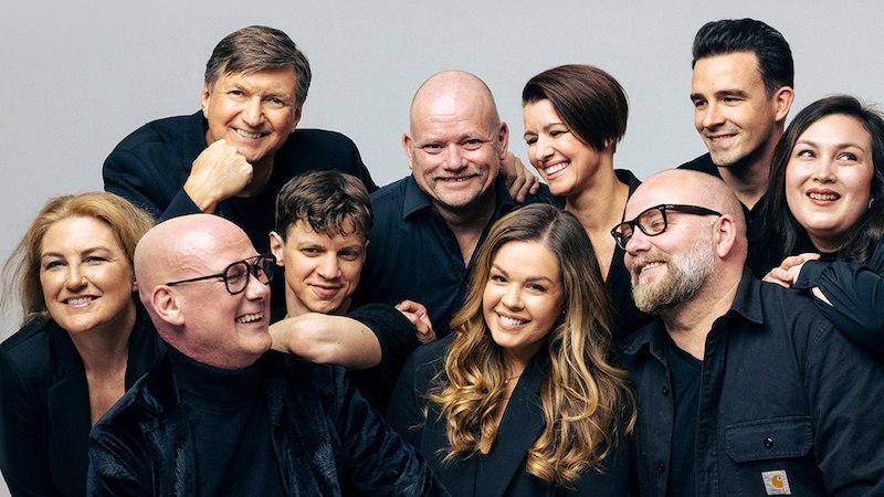 Ten people in black clothing smile cheerfully into the camera. They stand close together in front of a neutral background., &copy; GCM Konzerte