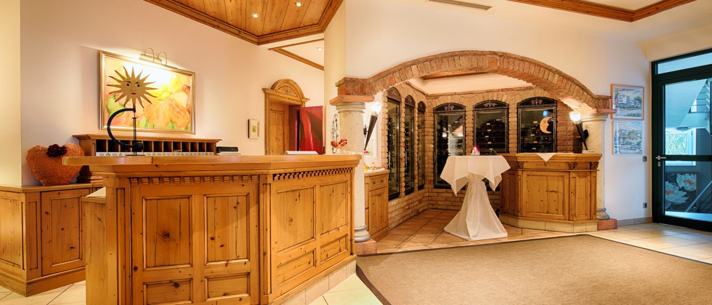 Rezeption im Hotel Landhaus Feckl mit Holzmöbeln, einem Gemälde, Weinregal und dekorativem Tisch. Gemütliche Atmosphäre mit Holzdecke und Beleuchtung., © Landhaus Franz Feckl