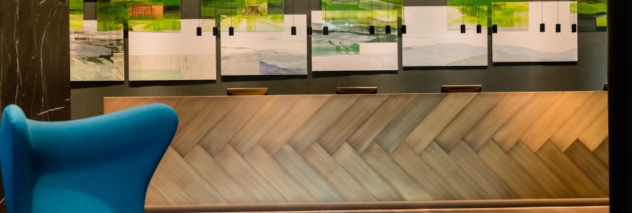 Moderne Rezeption mit h&ouml;lzerner Theke, blauer Sitzgelegenheit und Landschaftsbildern an der Wand. Elegantes, einladendes Ambiente., &copy; Motel One - Stuttgart Hauptbahnhof