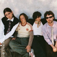 Vier M&auml;nner sitzen entspannt auf einer Bank, jeder mit einem individuellen Stil. Sie blicken in die Kamera, die Atmosph&auml;re wirkt l&auml;ssig und entspannt., &copy; links im Bild