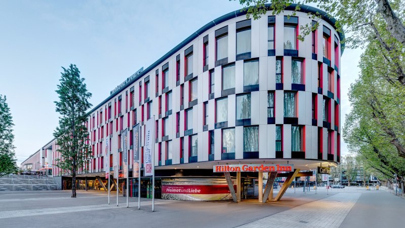 Modernes Hotelgebäude mit bunter Fassade, Hilton Garden Inn Stuttgart Neckarpark. Vor dem Gebäude stehen Fahnenmasten, umgeben von Bäumen., © Hilton Garden Inn Stuttgart Neckarpark