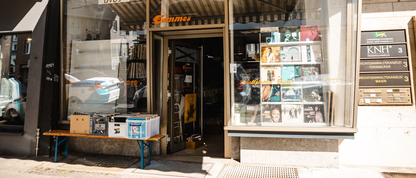 Plattenladen mit Schallplatten im Schaufenster, Tisch mit Kisten davor. Schild: "Tommes". Rechts ein Gebäude mit Kanzleischildern., © SMG Stuttgart Marketing GmbH - Sarah Schmid