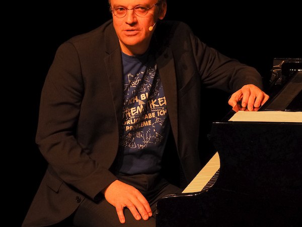 A man sits at a grand piano on a stage, wearing a microphone and a T-shirt with a mathematical print., &copy; Theaterhaus Stuttgart e.V.