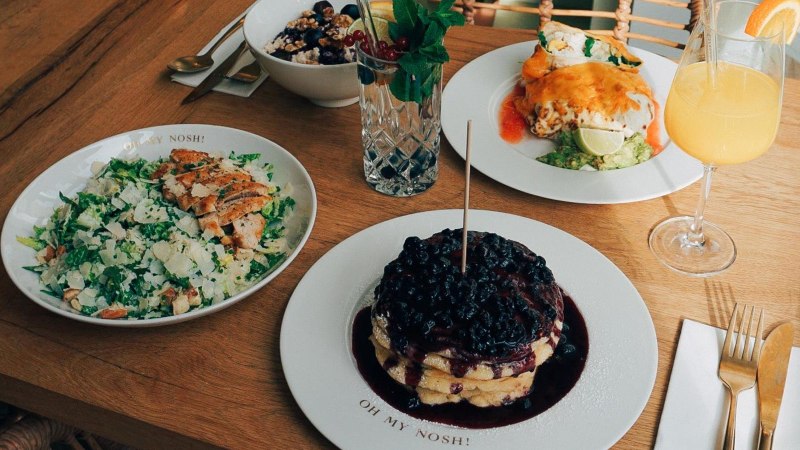 Ein Tisch mit Salat, Pfannkuchen mit Beeren, einem Getränk und weiteren Speisen auf Holztisch., © The Gardeners Nosh, Stuttgart