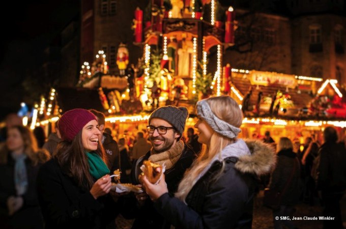 Drei Personen lachen und essen auf einem festlich beleuchteten Weihnachtsmarkt in Stuttgart. Im Hintergrund sind dekorierte Stände zu sehen., © Stuttgart Marketing GmbH