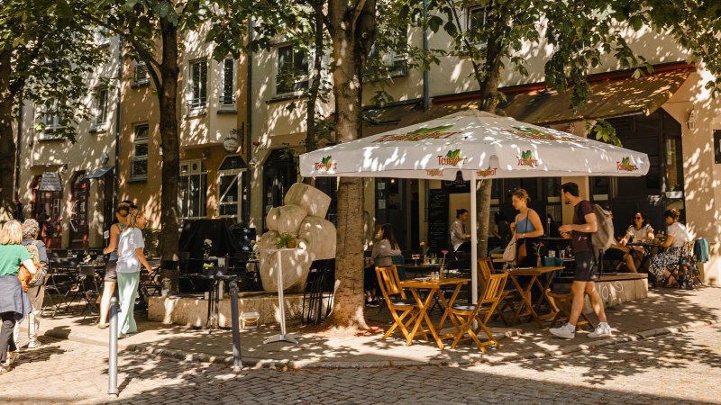 Ein gemütliches Straßencafé mit Sonnenschirm und Holztischen. Menschen sitzen und gehen vorbei, umgeben von Bäumen und Schatten., © Stuttgart Marketing GmbH - Sarah Schmid