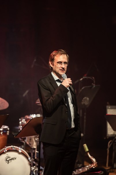 A man in a suit speaks into a microphone on a stage. Musical instruments such as a drum kit and music stands can be seen in the background., &copy; Staatliche Hochschule f&uuml;r Musik und Darstellende Kunst