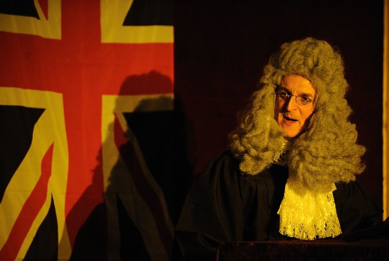 A man in a historic judge's robe and wig speaks in front of a British flag. The scene is bathed in warm light., © Gruseldinner GmbH & Co. KG A man in a historic judge's robe and wig speaks in front of a British flag. The scene is bathed in warm light., © Gruseldinner GmbH & Co. KG