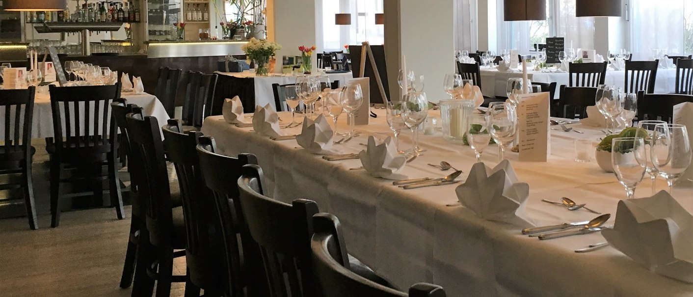 A stylishly decorated restaurant with long tables, white tablecloths, and dark wooden chairs. Subdued lighting and a bar in the background., &copy; bonjour Hotelbetriebs GmbH
