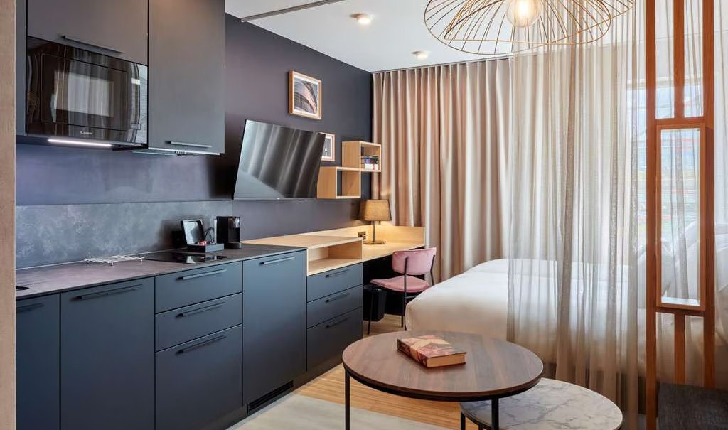 Modernes Studio mit dunkler Küche, Schreibtisch, Bett und stilvoller Beleuchtung. Vorhänge trennen den Schlafbereich ab., © Adagio