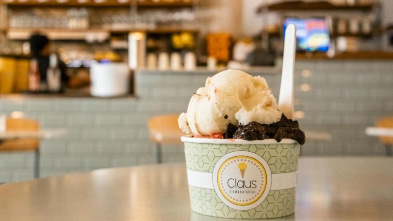 Ein Becher Eiscreme von Claus Eismanufaktur steht auf einem Tisch in einem Café. Der Hintergrund ist unscharf und zeigt eine Theke., © SMG, Sarah Schmid Ein Becher Eiscreme von Claus Eismanufaktur steht auf einem Tisch in einem Café. Der Hintergrund ist unscharf und zeigt eine Theke., © SMG, Sarah Schmid