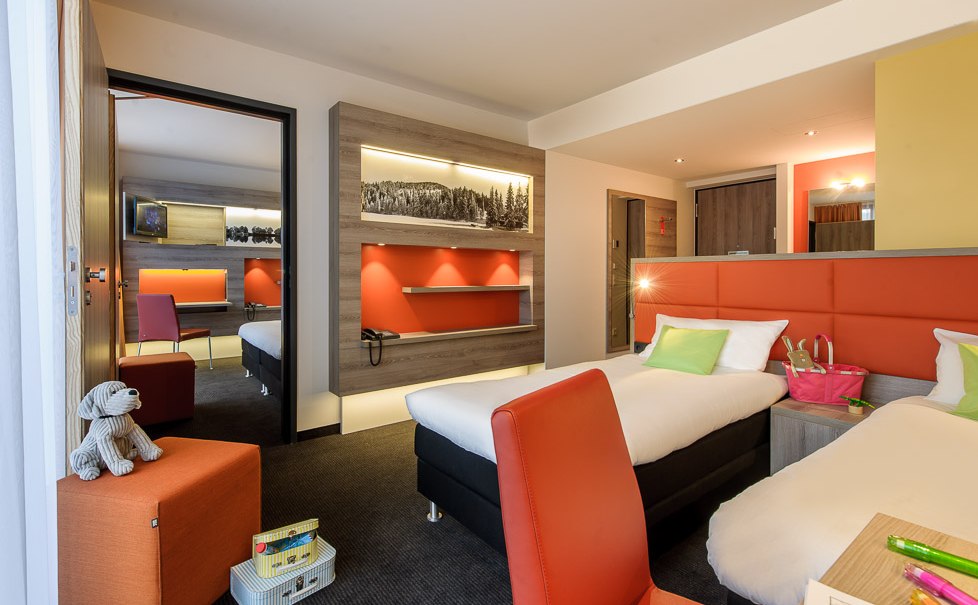 Modernes Hotelzimmer mit zwei Einzelbetten, roten Akzenten und einem angrenzenden Raum. Ein Plüschtier und Spielzeug sind auf einem Hocker platziert., © TOMAS Modernes Hotelzimmer mit zwei Einzelbetten, roten Akzenten und einem angrenzenden Raum. Ein Plüschtier und Spielzeug sind auf einem Hocker platziert., © TOMAS