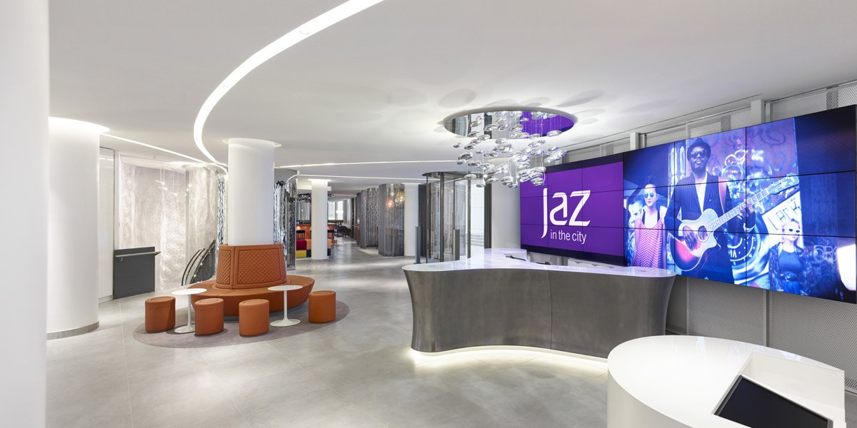 Modern reception with curved counter, large screen with "Jaz in the City" logo and stylish lighting., © Jaz Hotel Stuttgart Modern reception with curved counter, large screen with "Jaz in the City" logo and stylish lighting., © Jaz Hotel Stuttgart