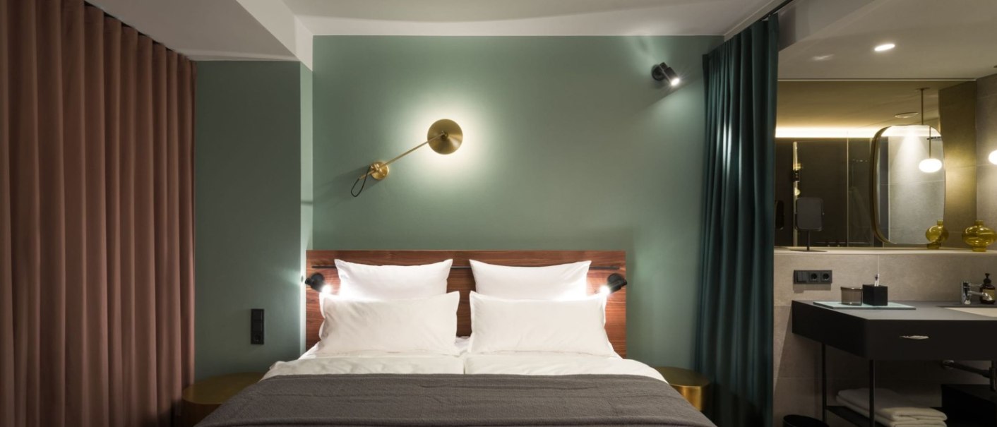 Modernes Hotelzimmer mit Doppelbett, grüner Wand, stilvollen Lampen und angrenzendem Badezimmer mit Waschbecken und Spiegel., © EmiLu Hotel GmbH