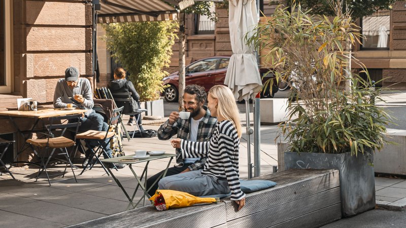 People enjoying coffee in front of a caf&eacute; in the sun. A couple is sitting on a bench, others at tables. Plants and parasols decorate the scene., &copy; SMG, Sarah Schmid
