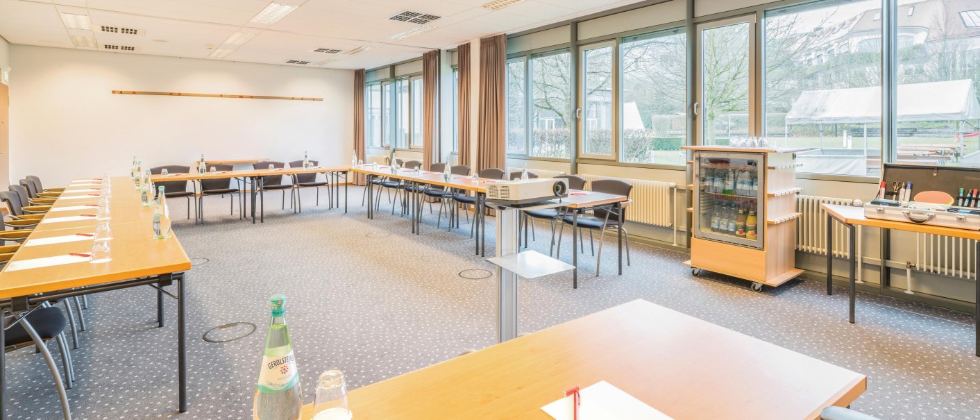 Heller Tagungsraum mit U-förmiger Tischanordnung, Stühlen, Projektor und großen Fenstern. Auf den Tischen stehen Wasserflaschen und Schreibmaterialien., © Seminaris Hotel Bad Boll