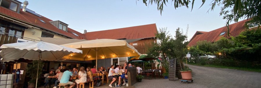 Menschen sitzen unter großen Sonnenschirmen in einem gemütlichen Hof mit roten Dächern und Bäumen., © Weinhof Zaiß Menschen sitzen unter großen Sonnenschirmen in einem gemütlichen Hof mit roten Dächern und Bäumen., © Weinhof Zaiß