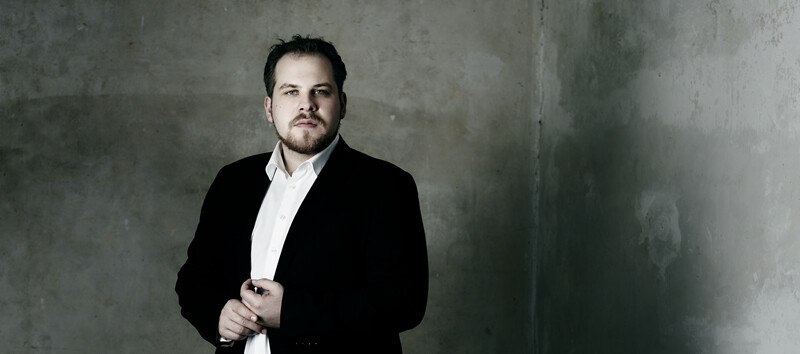 Ein Mann in einem schwarzen Anzug steht vor einer grauen, strukturierten Wand und blickt ernst in die Kamera., © Württembergische Staatstheater Stuttgart