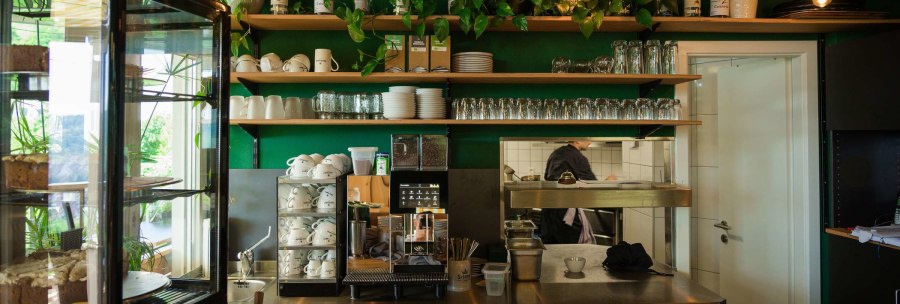 Café interior with coffee machine, cup shelves and cakes in a display case. Green walls and plants create a cozy atmosphere., © Rotenberger Weingärtle