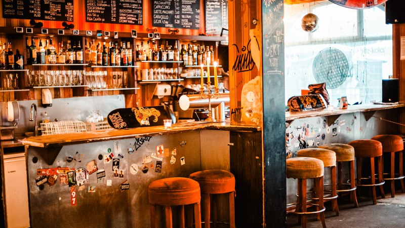 Gemütliche Bar mit Barhockern, einer Theke voller Flaschen und Gläser. Die Wände sind mit Stickern dekoriert, und eine Tafel zeigt Getränkepreise., © Mata Hari Gemütliche Bar mit Barhockern, einer Theke voller Flaschen und Gläser. Die Wände sind mit Stickern dekoriert, und eine Tafel zeigt Getränkepreise., © Mata Hari