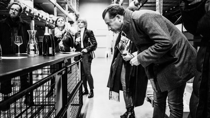 Mehrere Personen stehen in einem Lagerraum und probieren Champagner. Eine Person beugt sich vor, um ein Glas zu riechen. Schwarz-Weiß-Foto., © Bernhard Kahrmann