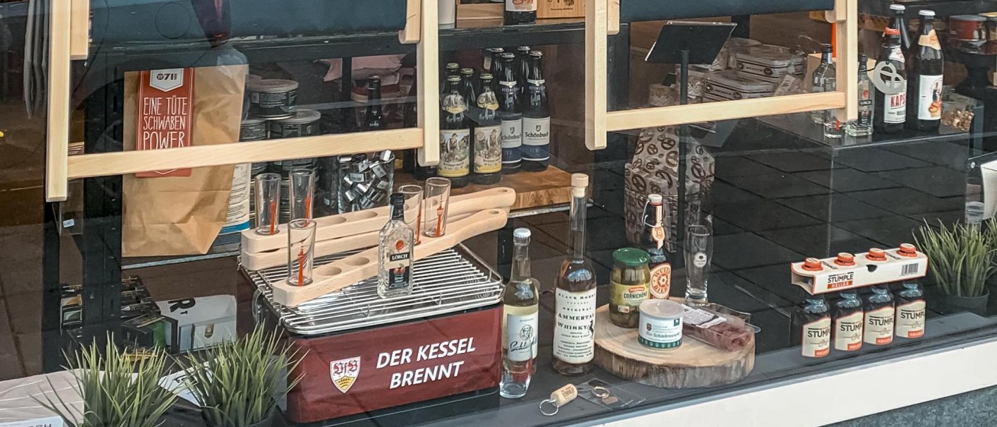 Schaufenster mit regionalen Produkten wie Bier, Spirituosen und Lebensmitteln. Dekoriert mit Pflanzen und Werbeschildern. Schriftzug 'Der Kessel brennt' sichtbar., © 0711 Store, Stuttgart