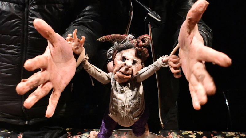 A grotesque doll with a distorted face is held up by two large hands. The doll is wearing a shirt and has striking facial features., © FITZ Das Theater animierter Formen A grotesque doll with a distorted face is held up by two large hands. The doll is wearing a shirt and has striking facial features., © FITZ Das Theater animierter Formen