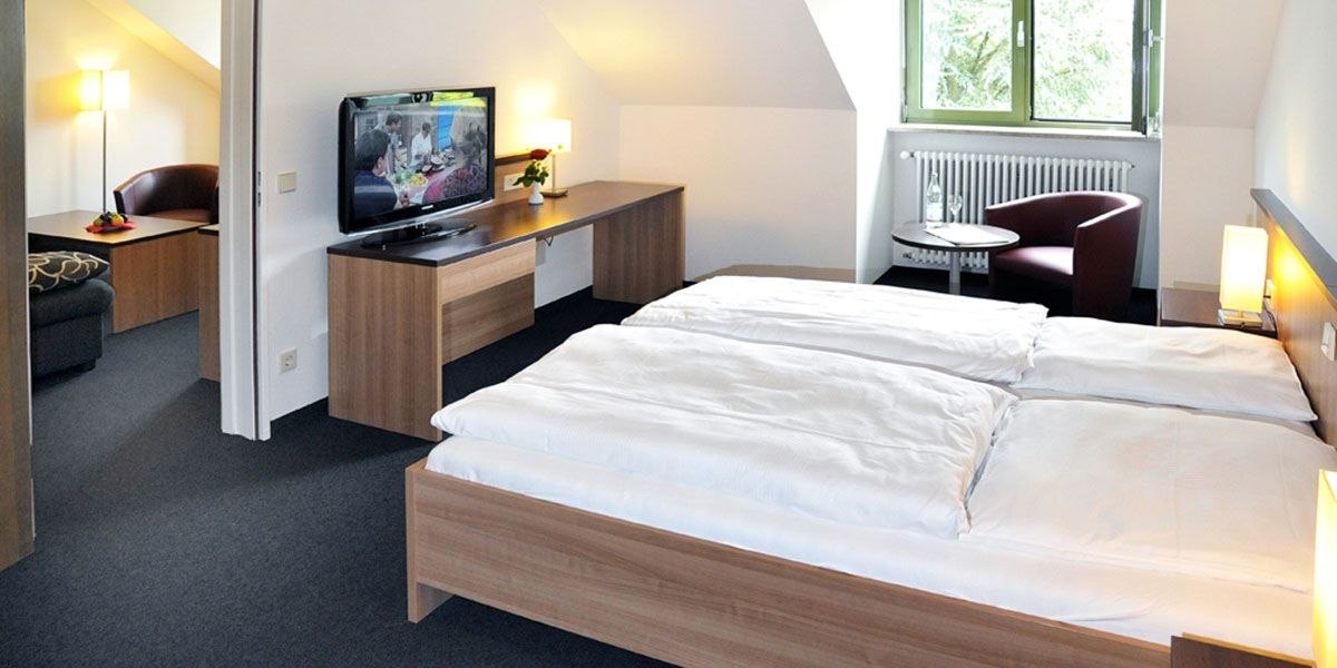 Modernes Doppelzimmer mit Doppelbett, Schreibtisch, Fernseher und Sitzbereich. Helle Einrichtung und Fenster mit Blick ins Grüne., © Christkönigshaus