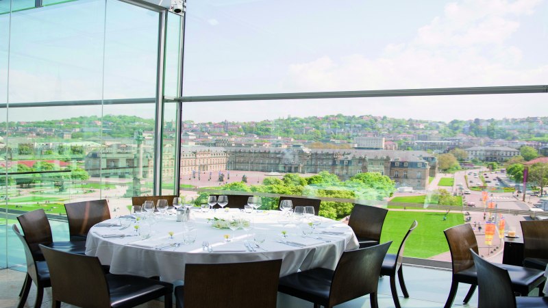 Ein runder Tisch mit weißer Tischdecke und Gläsern steht in einem verglasten Raum mit Blick auf eine Stadt und historische Gebäude., © Stuttgart-Marketing GmbH Ein runder Tisch mit weißer Tischdecke und Gläsern steht in einem verglasten Raum mit Blick auf eine Stadt und historische Gebäude., © Stuttgart-Marketing GmbH