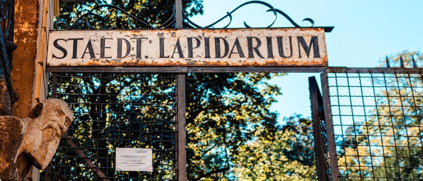 Eingang zum Städtischen Lapidarium mit einem alten, verwitterten Schild und einer Steinskulptur. Im Hintergrund sind Bäume zu sehen., © Stuttgart Marketing GmbH, Sarah Schmid
