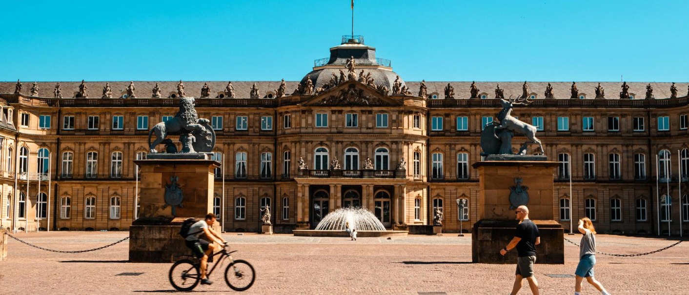 Das Neue Schloss Stuttgart bei sonnigem Wetter. Menschen gehen und ein Fahrradfahrer f&auml;hrt im Vordergrund. Zwei Statuen flankieren den Eingang., &copy; Stuttgart-Marketing GmbH, Sarah Schmid