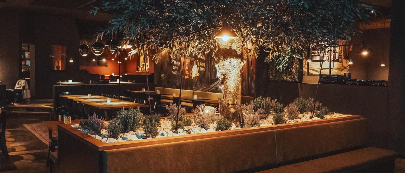 Warm beleuchteter Restaurantinnenraum mit großem künstlichen Baum als Mittelpunkt und modernen Sitzbereichen., © L'Artista Stuttgart GmbH Warm beleuchteter Restaurantinnenraum mit großem künstlichen Baum als Mittelpunkt und modernen Sitzbereichen., © L'Artista Stuttgart GmbH