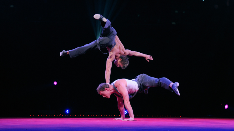 Zwei Artisten zeigen eine beeindruckende akrobatische Performance auf einer Zirkusbühne. Einer balanciert auf den Händen, während der andere auf dessen Hals balanciert., © Foto Thierry Bissat