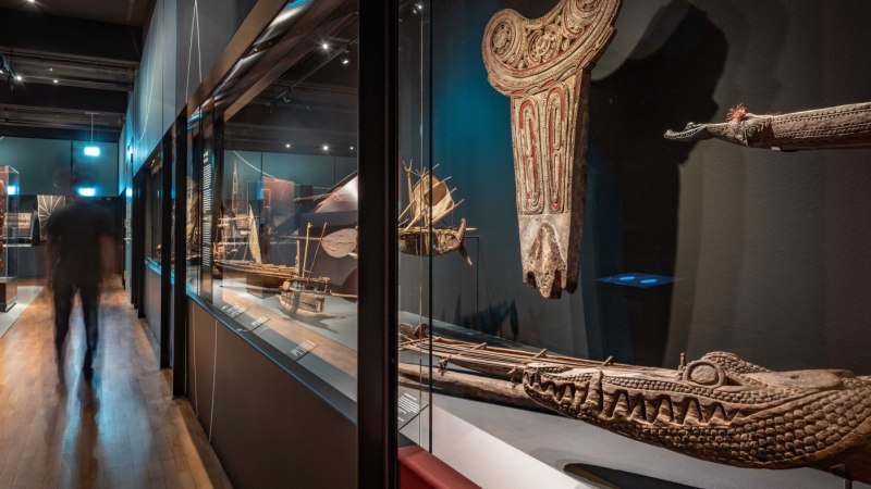 Ein Museumsraum mit ozeanischen Kunstwerken und Modellen von Booten in Glasvitrinen. Eine Person geht durch den Raum., © Linden-Museum Stuttgart