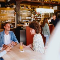 Three people sit laughing at a table in a cozy bar. Glasses and bottles can be seen in the background., © © Stuttgart-Marketing GmbH, Alwin Maigler Three people sit laughing at a table in a cozy bar. Glasses and bottles can be seen in the background., © © Stuttgart-Marketing GmbH, Alwin Maigler