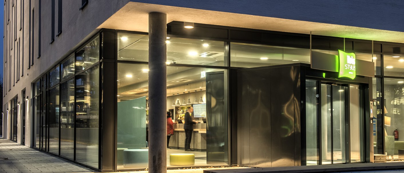 Modernes Hotelgebäude mit Glasfassade und beleuchtetem Eingang bei Nacht. Innen sind Personen an der Rezeption zu sehen., © ibis Styles Stuttgart Vaihingen
