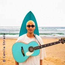 Eine Person mit Sonnenbrille hält eine türkisfarbene Gitarre vor einem Surfbrett am Strand., © links im Bild